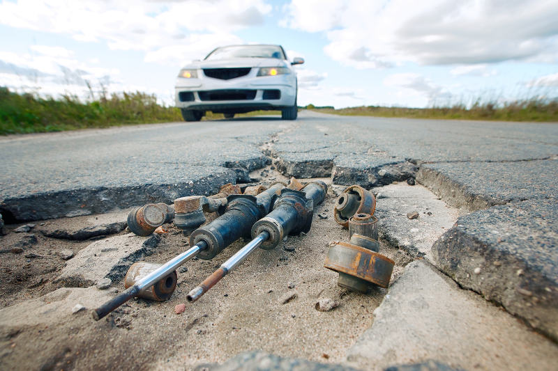 A broken suspension parts lying on a bumpy road, the car blurred in the background.